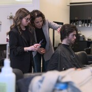 Une femme en train de donner des conseil à une étudiante derrière un homme assis sur une chaise de coiffure.