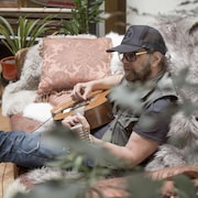 Un homme qui joue de la guitare, assis sur un sofa.