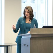 Femme vêtue de turquoise parle à un podium en pointant la salle. 9 mars 2023.