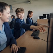 Des élèves qui sont devant des ordinateurs dans une salle de classe; l'un d'eux montre du doigt un écran.