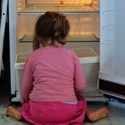 Une enfant est accroupie face à un réfrigérateur vide.