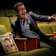 Un comédien est assis dans une salle de théâtre, avec un portrait photo déposé sur un siège à ses côtés.