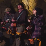 Trois musiciens vêtus d'équpiement de spéléologie jouent de la guitare dans une grotte. 