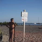 Un homme nu au bord de la plage. Un panneau indique que les vêtements y sont optionnels.