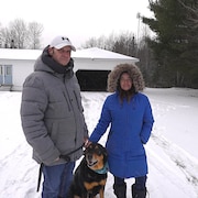 Un homme, sa femme et leur chien devant une maison.