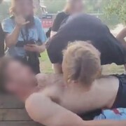 Un jeune homme couch sur une table  pique-nique torse nu a un shooter d'alcool sur la poitrine. Une autre personne boit sans utiliser ses mains.