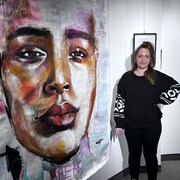 Une femme se tient debout dans une salle d'exposition, à côté d'une grande toile, où le visage d'une femme est représenté.