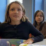 Une élève assise à son bureau. À sa gauche et derrière elle, il y a une autre fille et un garçon.