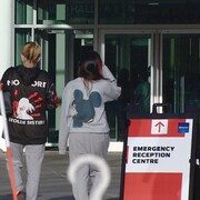 Trois femmes de dos marchand vers les portes vitrées d'un édifice. Un écriteau rouge et blanc avec la mention emergency reception centre. 5 mai 2023.