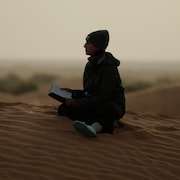 Une femme est assise par terre dans un désert. Elle tient un cahier en mains et regarde au loin.