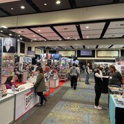 Plan large sur les stands du Salon du livre de l'Outaouais. Des auteurs sont attablés. Des passants circulent.