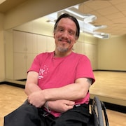 Un homme en fauteuil roulant sourit dans un studio de danse. Son reflet apparaît dans le miroir derrière lui.