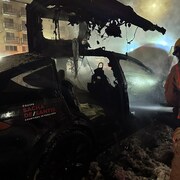 Une voiture extrêmement endommagée par les flammes. Un pompier essaie de l'éteindre.  