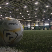 En avant plan un balon de soccer déposé sur un gazon artificiel dans un centre d'entraînement. En fond, des joueuses qui s'entraînent. Mars 2025.
