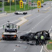 Une zone délimitée par un ruban adhésif montre une voiture endommagée et des véhicules de police garés à côté.