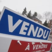 À l'extérieur,  dans une rue résidentielle, gros plan sur une pancarte  indiquant « Vendu » et qui surmonte une autre pancarte marquée  « À vendre ».