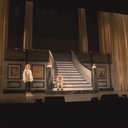 Quatre personnes sur une scène avec un escalier de marbre et de long rideaux blancs.