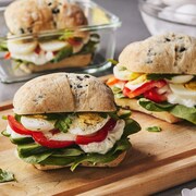 Trois sandwichs aux œufs à la grecque sur une planche de bois avec des plats pour les lunchs.