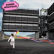 Une fille joue avec un cerf-volant multicolore devant un usine avec beaucoup de ventilateurs, à côté du logo de la bonne nouvelle de MAJ.