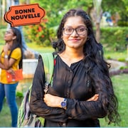 Une femme indienne avec un sac à dos sourit dans un jardin devant deux autres étudiantes, à côté du logo « Bonne nouvelle » de MAJ.
