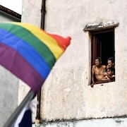 Trois personnes regardent à travers une fenêtre, à côté d'un drapeau arc-en-ciel.