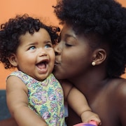 La fillette a la peau métisse et des frisettes, la maman a la peau noire et une coupe afro courte. La maman fait un bisou à son enfant qui sourit. 