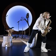 On voit un personnage jouer du saxophone. Il porte des bottes et une veste avec des fausses mains crochetées. 
