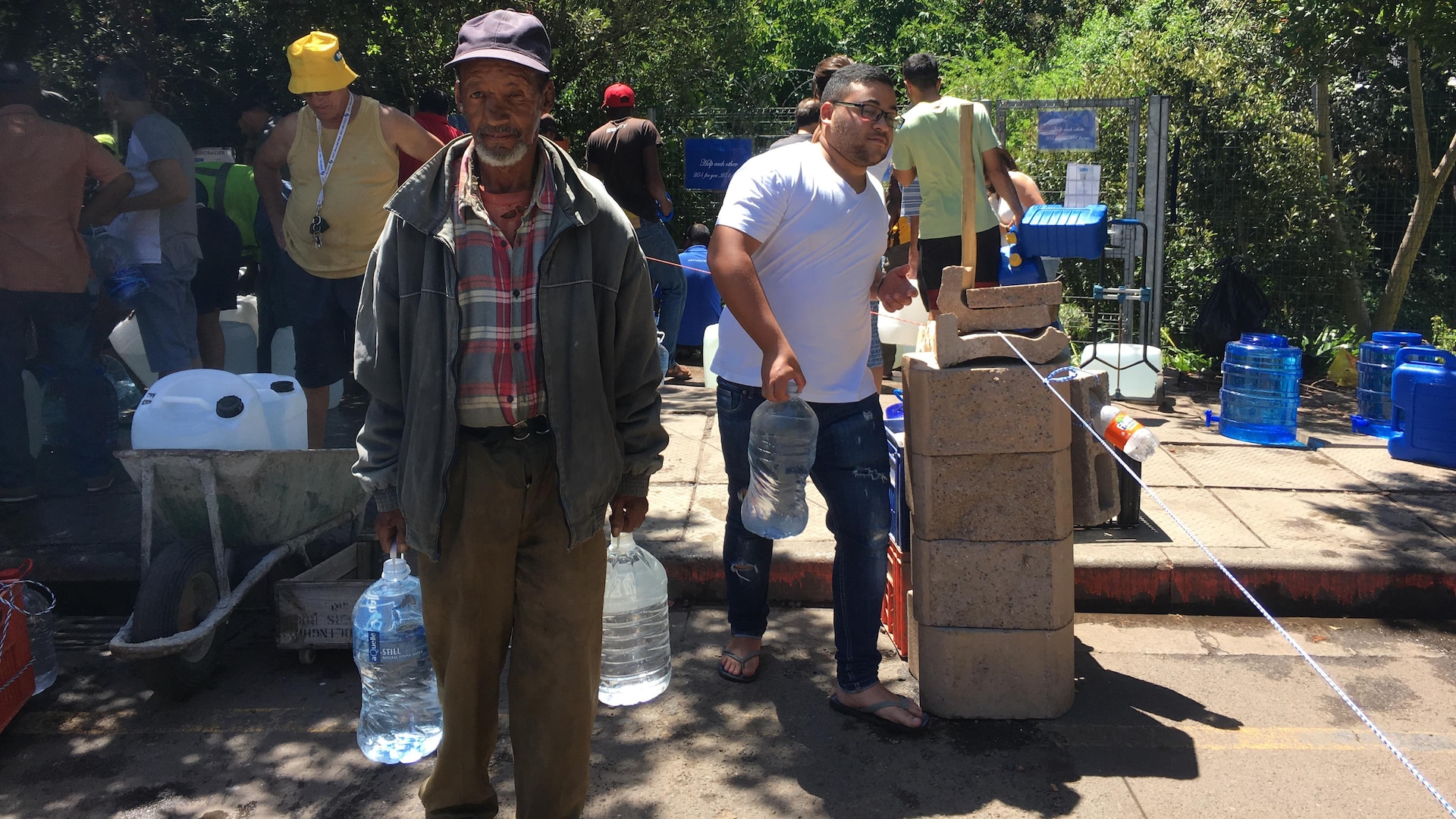 Un homme âgé transporte deux bidons d'eau. En arrière-plan, d'autres Capetoniens vont remplir leurs bidons.