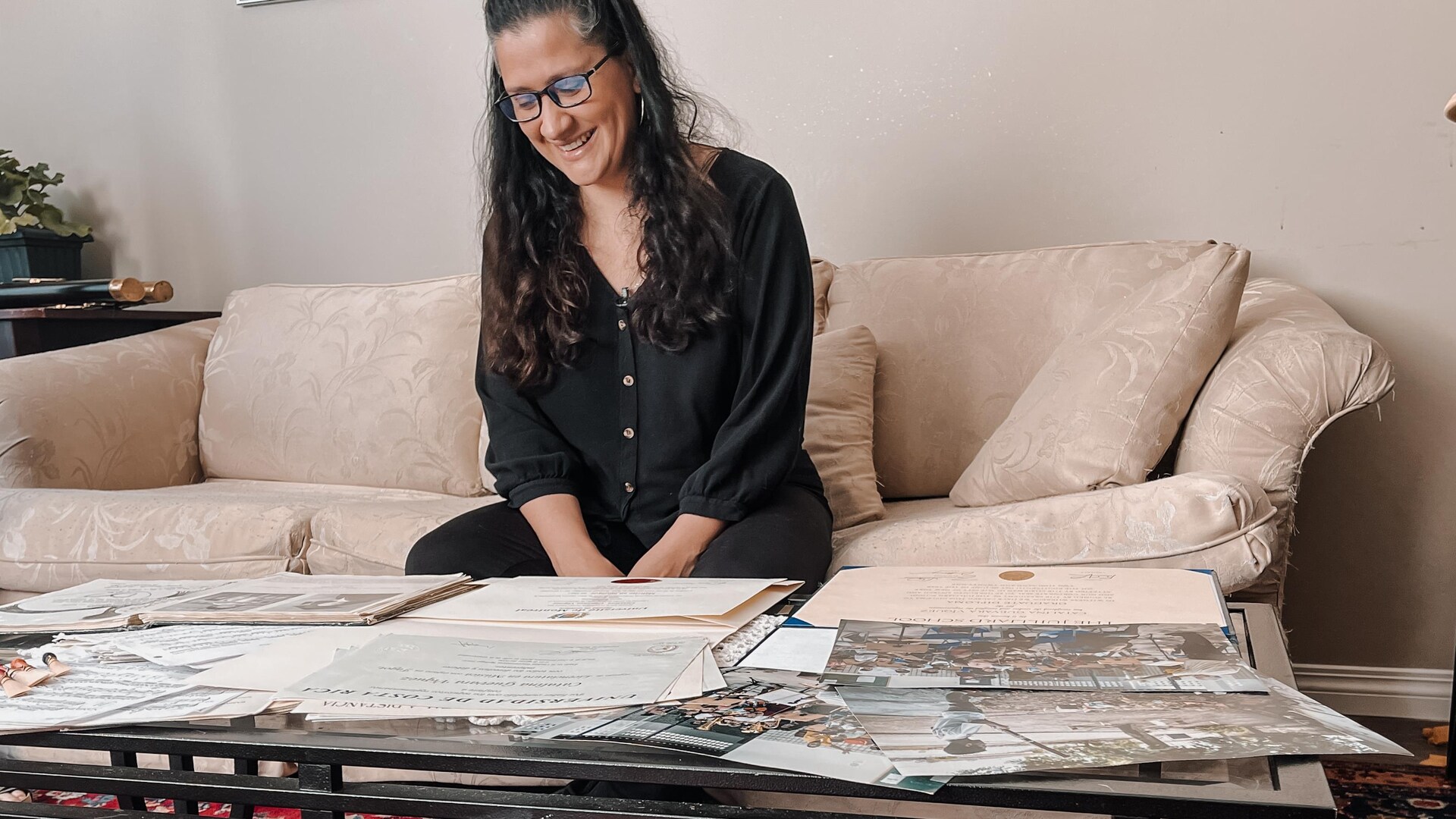 Catalina Klein assise sur un sofa regarde des certificats et des photos déposés sur une table de salon. 