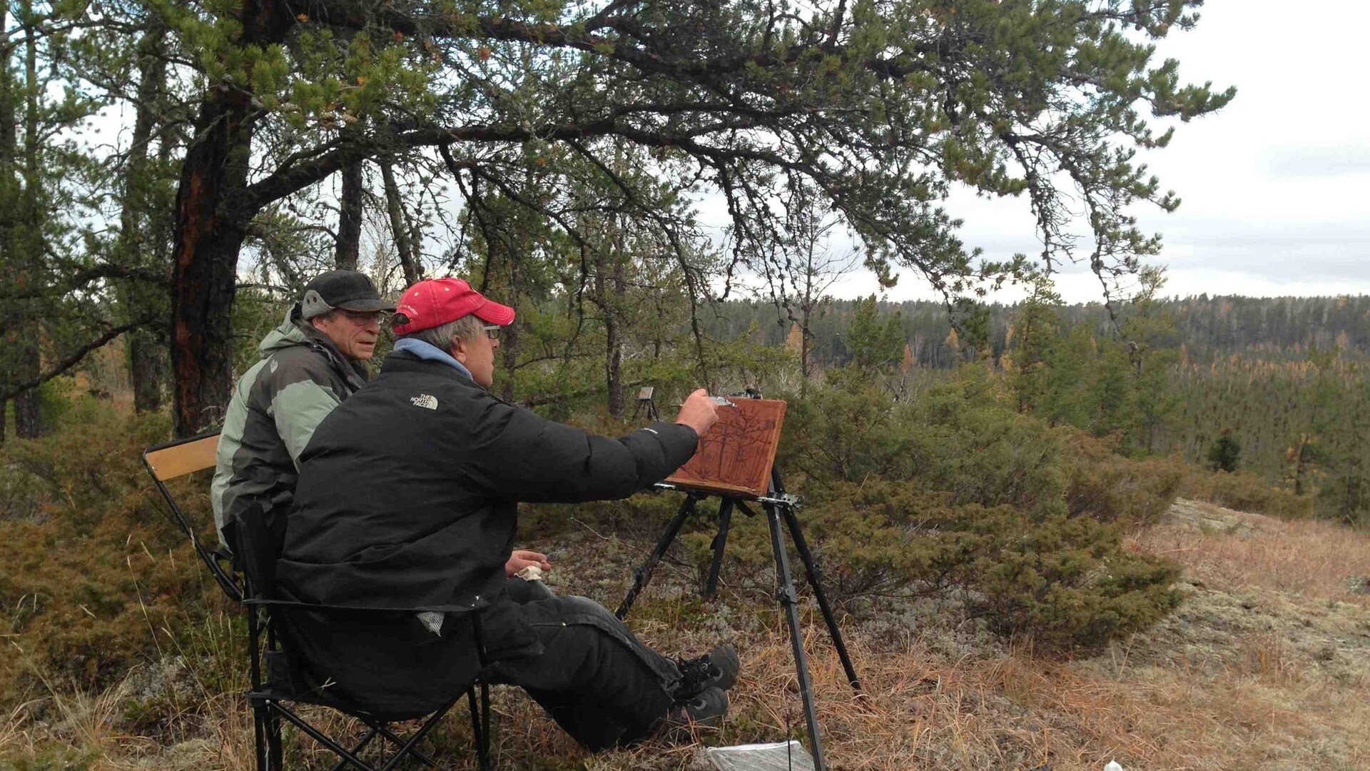 David MacNair faisant une esquisse devant un paysage. Son ami Jim est assis à ses côtés.