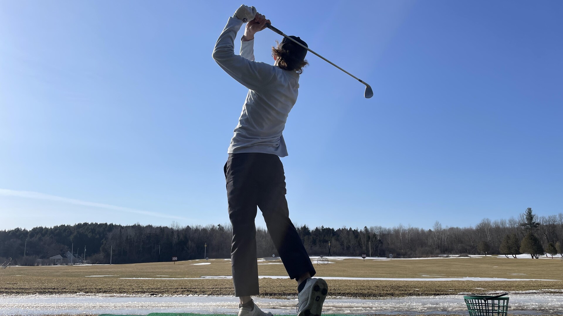 A golfer practices on a partially snow-covered driving range.