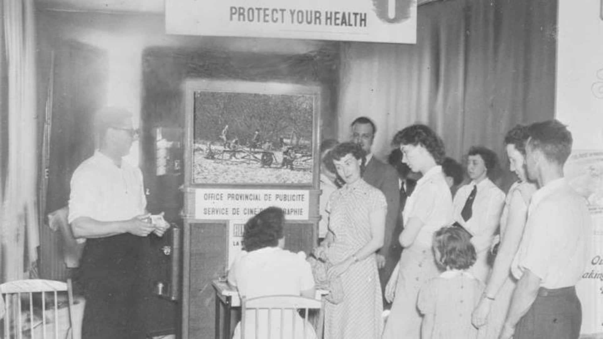 Le personnel infirmer au travail auprès du grand public lors d'un atelier de prévention contre la tuberculose en 1949. Des gens regardent un petit film pendant qu'on fait des radios pulmonaires à d'autres derrière un rideau.