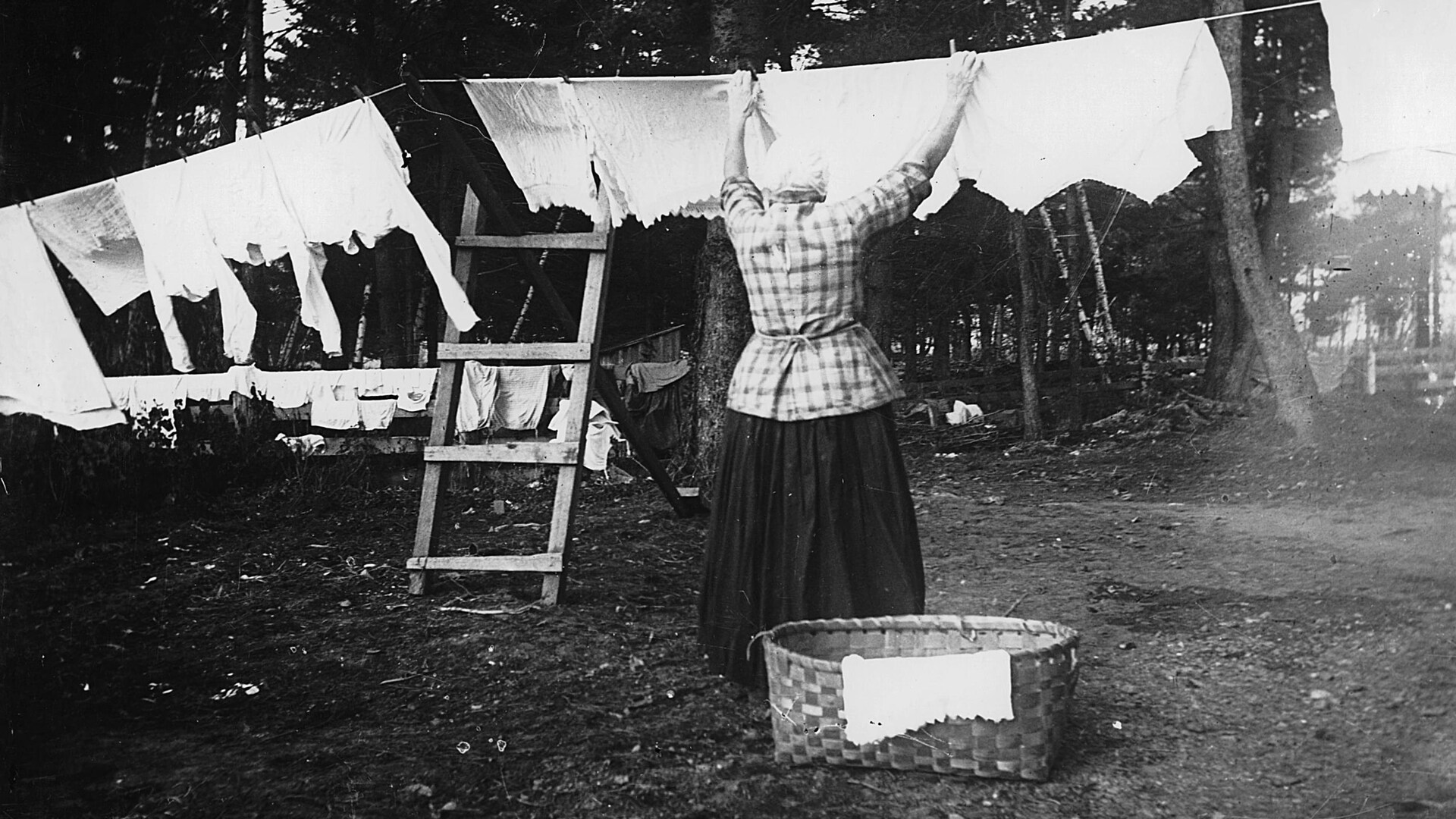 Une femme de dos est occupée à suspendre le linge de toute la famille sur une corde à linge, au milieu d'arbres, sur une photographie du début du siècle en noir et blanc. La lessive est immaculée. 