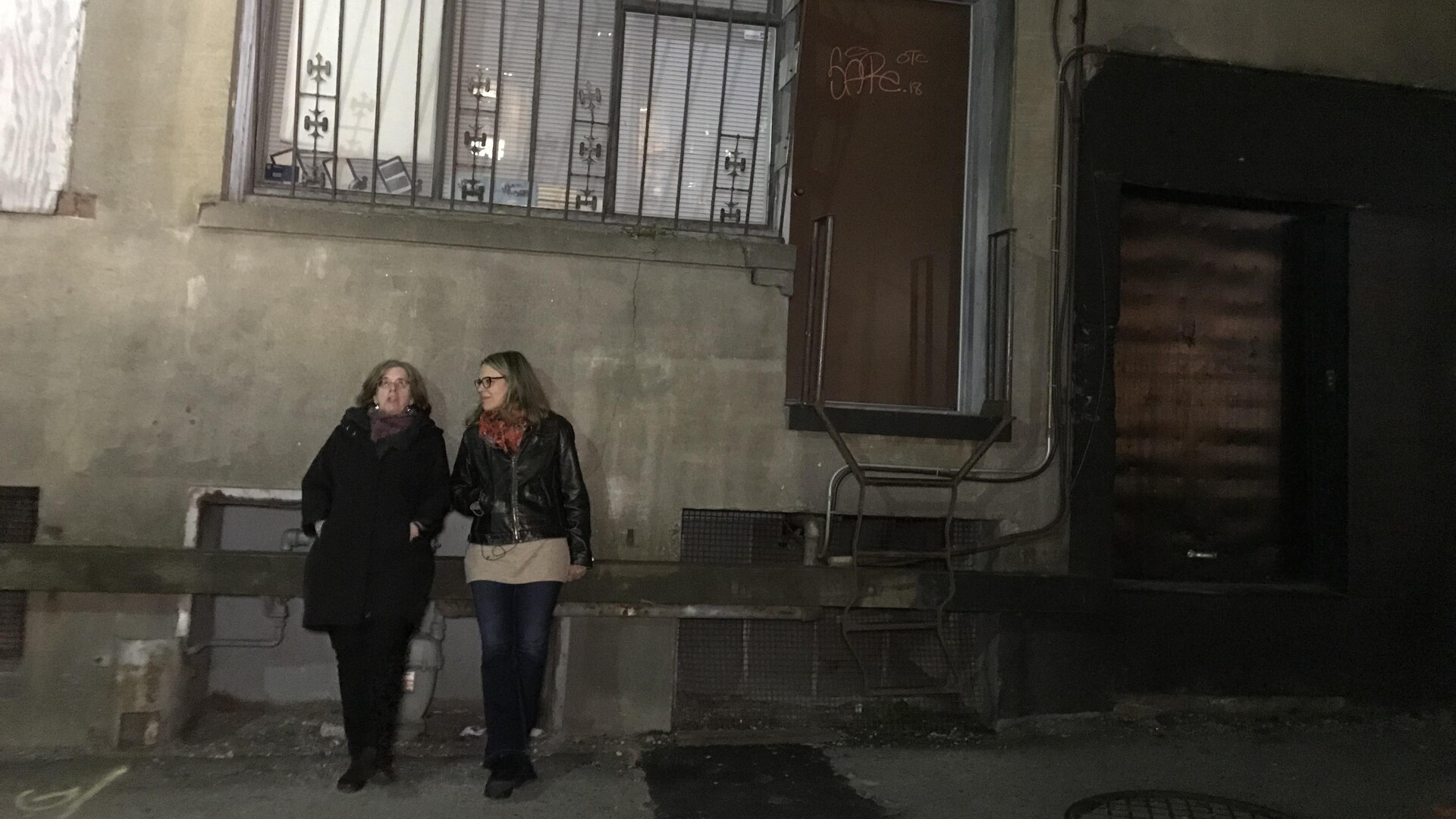 Deux femmes adossées sur le mur d'un immeuble dans une ruelle, sous une fenêtre grillagée éclairée.