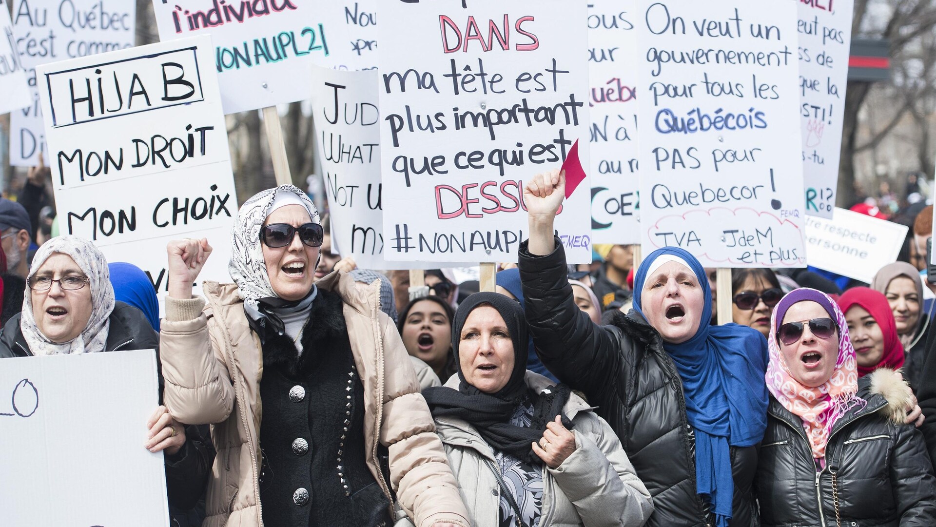 Des femmes voilées manifestent avec des pancartes disant entre autres « Hidjab, mon droit mon choix », « Ce qui est dans ma tête est plus important que ce qui est dessus », « On veut un gouvernement pour tous les Québécois, pas pour Québecor! »