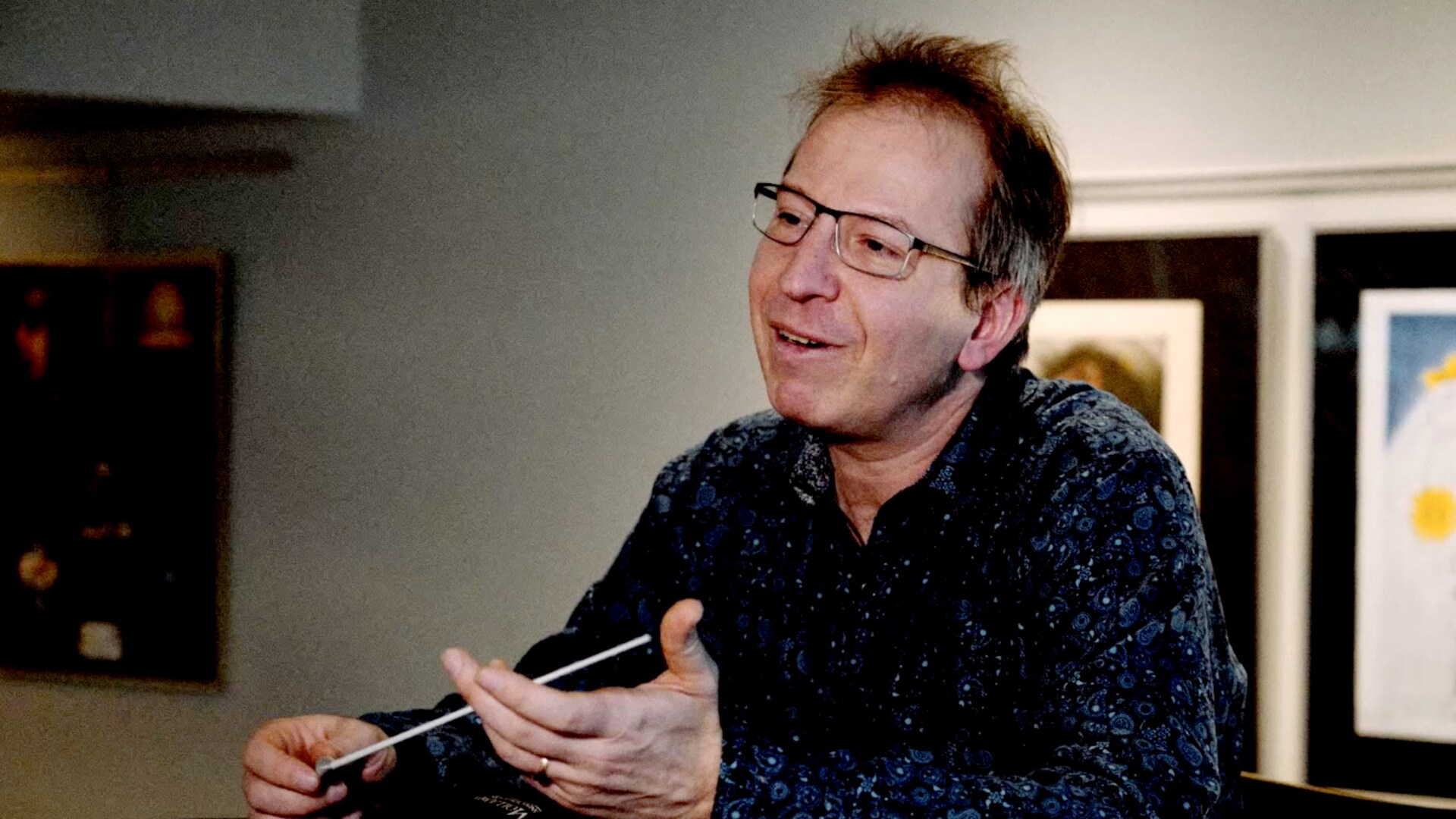 Pierre Simard, directeur artistique et musical du Vancouver Island Symphony assis en discutant, tenant de sa main droite la baguette du chef d'orchestre.
