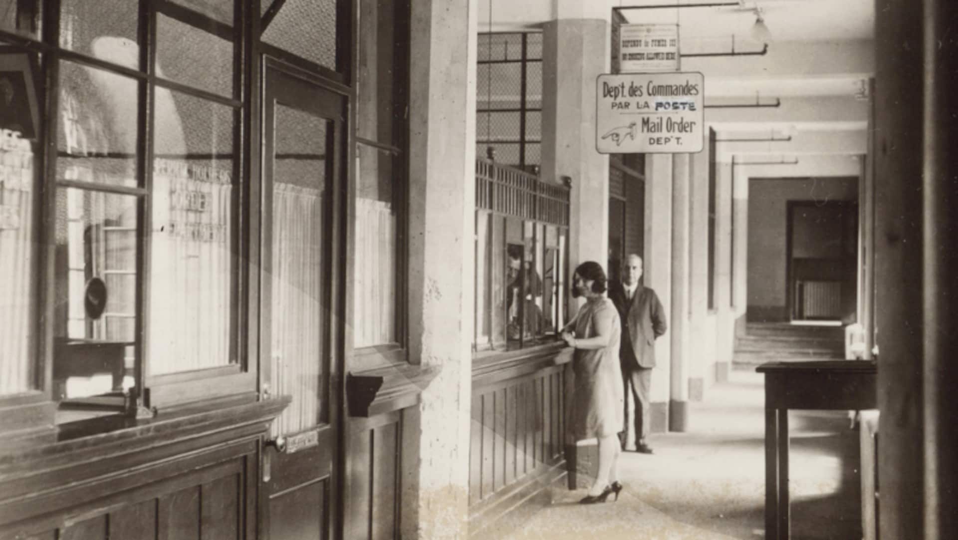 On aperçoit une femme à un comptoir, au bout d'un corridor. Une affiche indique qu'il s'agit du département des commandes par la poste. 