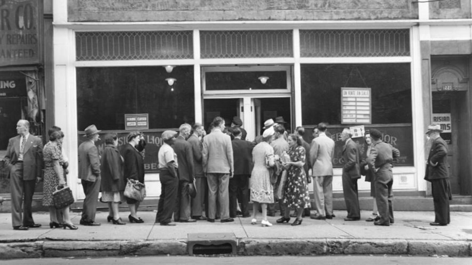Une foule de clients se pressent devant une succursale de la Commission des liqueurs, en 1945. La file part des deux côtés, et il y a beaucoup d'animation. Plusieurs clients - il y autant d'hommes que de femmes - fument d'un air résigné. Un panneau annonçant des prix en solde est affiché en vitrine, mais on ne voit rien à l'intérieur. Une foule de clients se pressent devant une succursale de la Commission des liqueurs, en 1945. La file part des deux côtés, et il y a beaucoup d'animation. Plusieurs clients - il y autant d'hommes que de femmes - fument d'un air résigné. Un panneau annonçant des prix en solde est affiché en vitrine, mais on ne voit rien à l'intérieur.