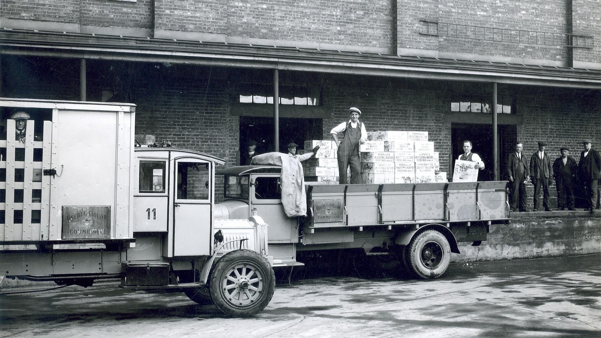 Des employés de la Commission des liqueurs sortent des caisses des entrepôts pour les charger dans les camions de livraison. L'un d'entre eux sourit à la caméra, pendant que d'autres surveillent des opérations à partir des quais. Des employés de la Commission des liqueurs sortent des caisses des entrepôts pour les charger dans les camions de livraison. L'un d'entre eux sourit à la caméra, pendant que d'autres surveillent des opérations à partir des quais.