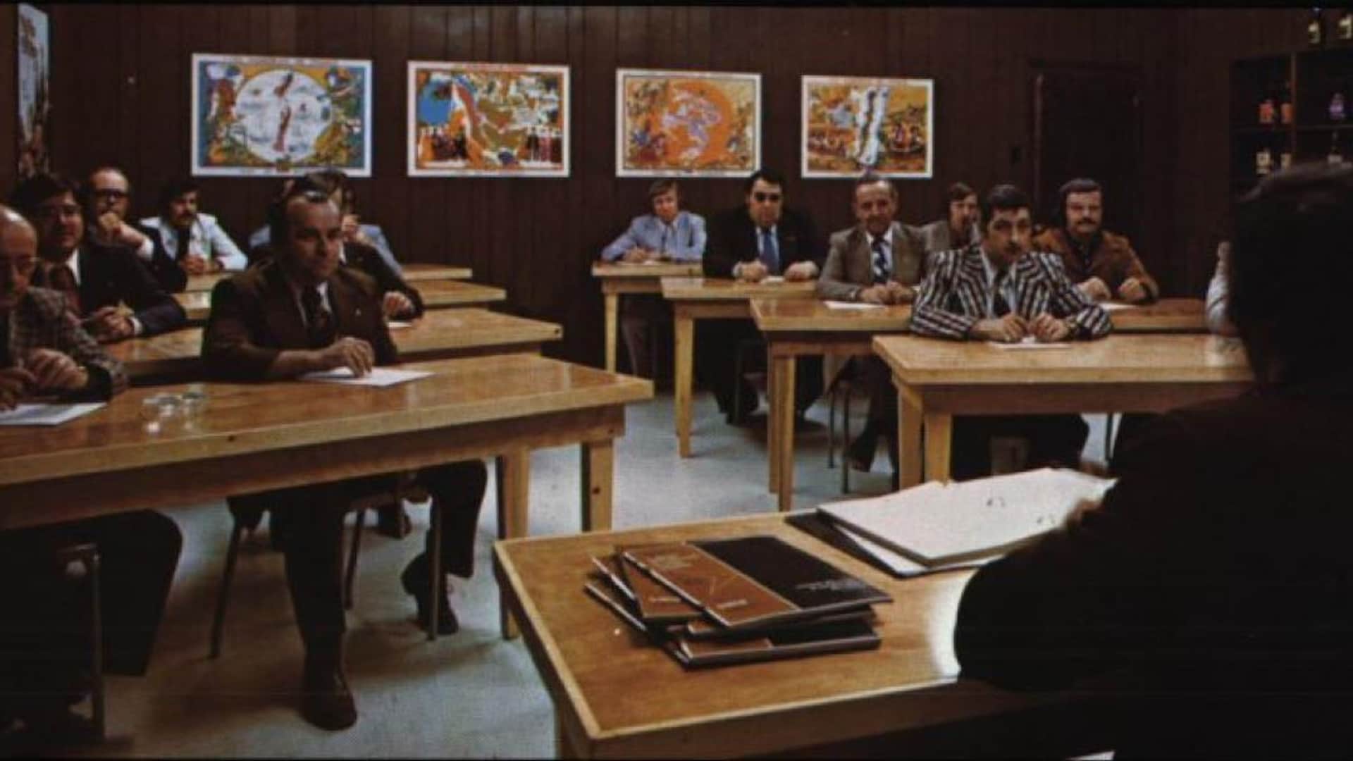 Des hommes écoutent un professeur, on les voit de face, ils sont tous en complet et en cravate, à la mode des années 1970. Au mur, on voit des bouteilles, et des cartes au fond. Les murs sont tous en lattes de bois foncé. 