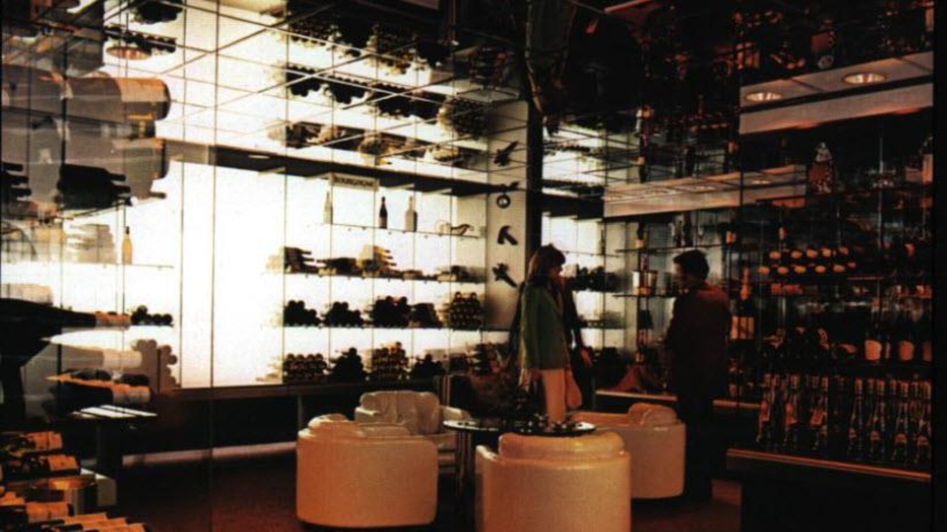 Deux personnes en discussions dans un espace aéré, lumineux et moderne, au milieu d'étagères esthétiquement chargées de bouteilles.