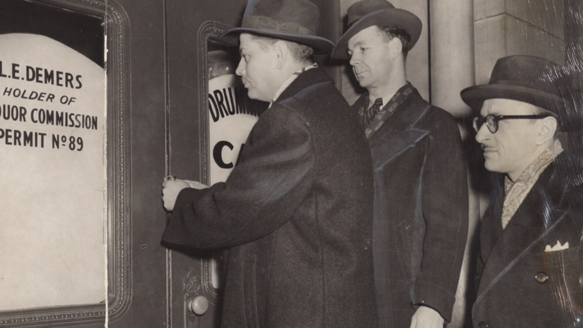 Trois hommes se tiennent devant la porte d'un club de nuit. On peut lire le nom du propriétaire (L.E. Demers) et le numéro de son permis (89) affiché dans la porte. Les hommes de la Police des liqueurs sont en civil. Trois hommes se tiennent devant la porte d'un club de nuit. On peut lire le nom du propriétaire (L.E. Demers) et le numéro de son permis (89) affiché dans la porte. Les hommes de la Police des liqueurs sont en civil.