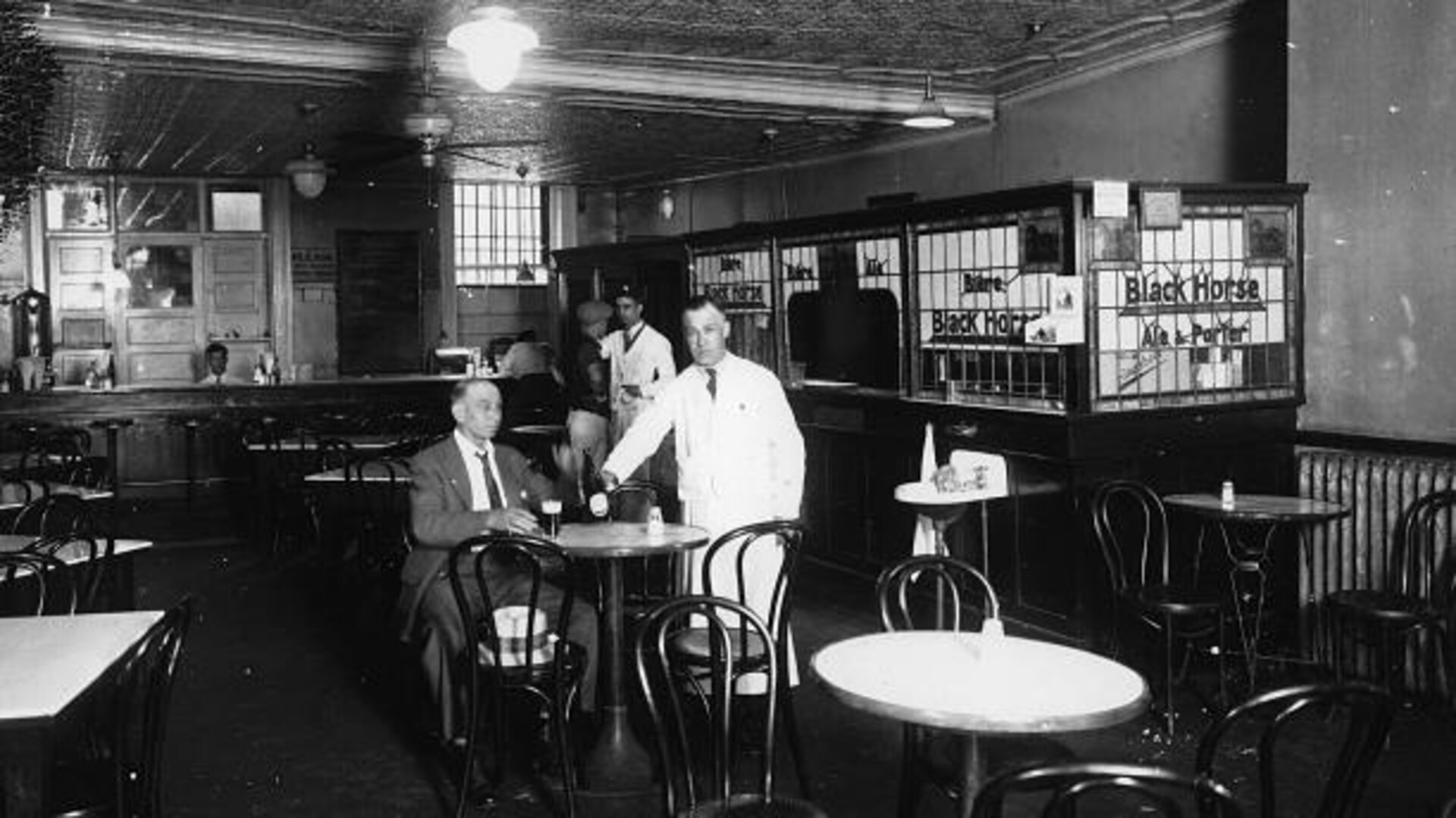 Un client esseulé est installé devant une pinte de bière dans une taverne du début du 20e siècle. Il est le seul client de l'endroit, près de lui, le serveur en sarrau blanc tient une bouteille de bière. Autour d'eux, toutes les tables sont vides. Un client esseulé est installé devant une pinte de bière dans une taverne du début du 20e siècle. Il est le seul client de l'endroit, près de lui, le serveur en sarrau blanc tient une bouteille de bière. Autour d'eux, toutes les tables sont vides.