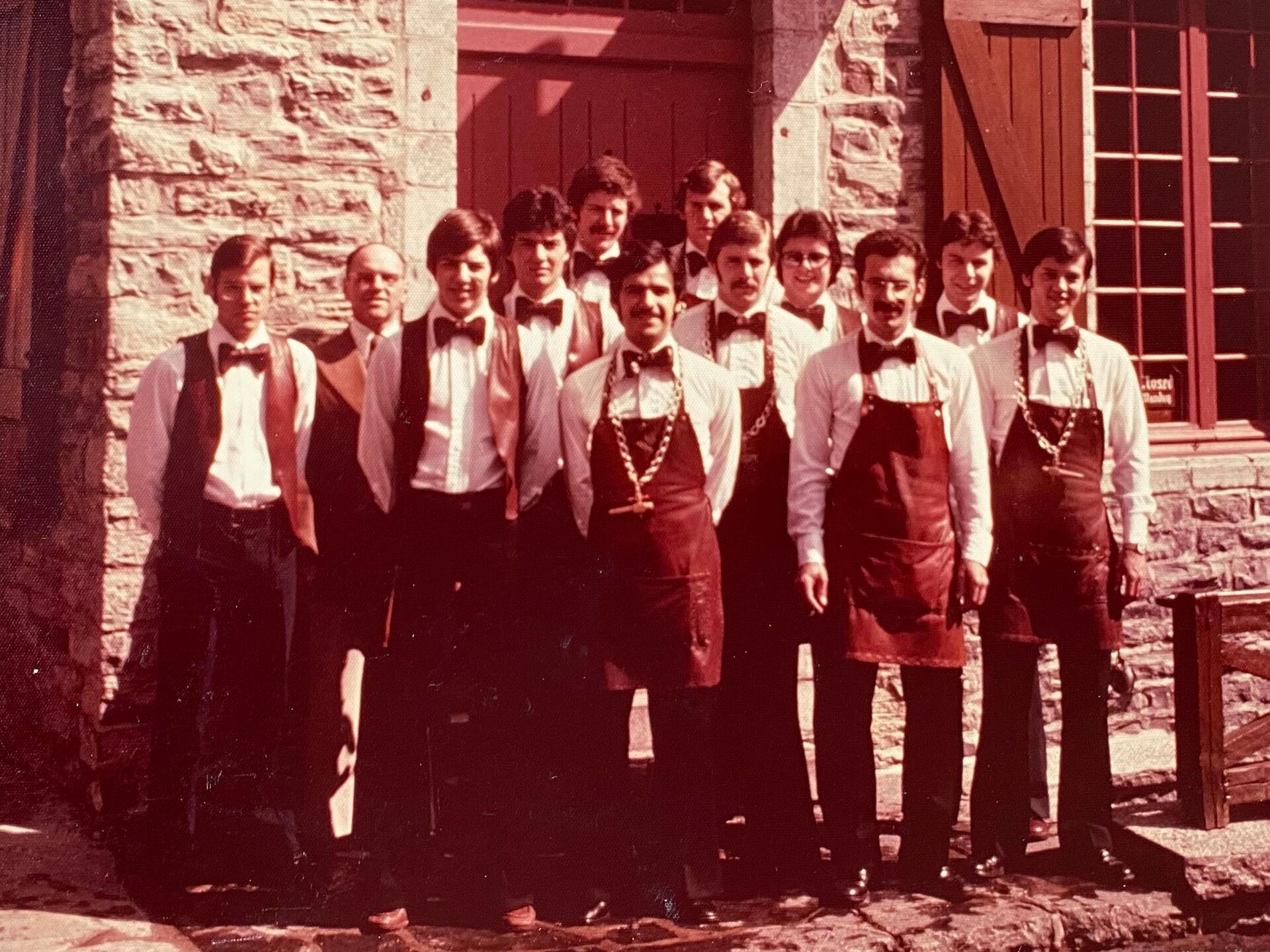 Un groupe souriant de conseillers en vin et de commis de la SAQ arborent noeuds papillon, tabliers de cuirs et petites vestes devant une maison patrimoniale, dans les années 1970. Il s'agit de la première Maison des vins du réseau, sur la Place-Royale, 