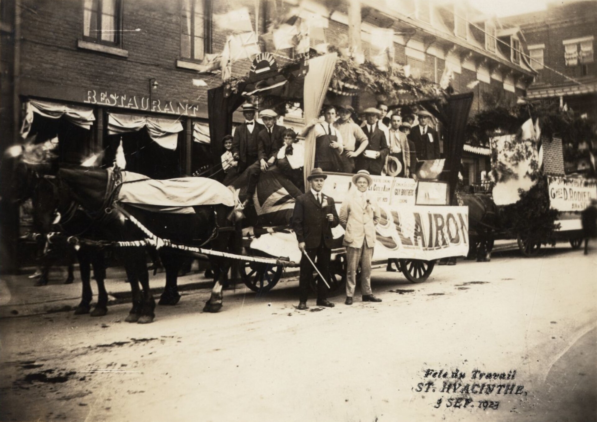 Un défilé de la fête du travail en 1923, à Saint-Hyacinthe. On voit des hommes montés sur le char du journal Le Clairon tiré par deux chevaux, et un char autre abondamment décoré derrière. Un défilé de la fête du travail en 1923, à Saint-Hyacinthe. On voit des hommes montés sur le char du journal Le Clairon tiré par deux chevaux, et un char autre abondamment décoré derrière.