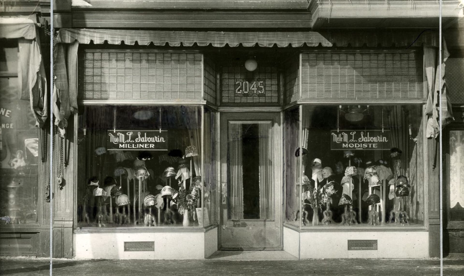 Vitrine d'une modiste du début du 20e siècle, à Montréal. On voit des dizaines de jolis chapeaux montés sur des présentoirs, de part et d'autre de la porte d'entrée. Vitrine d'une modiste du début du 20e siècle, à Montréal. On voit des dizaines de jolis chapeaux montés sur des présentoirs, de part et d'autre de la porte d'entrée.