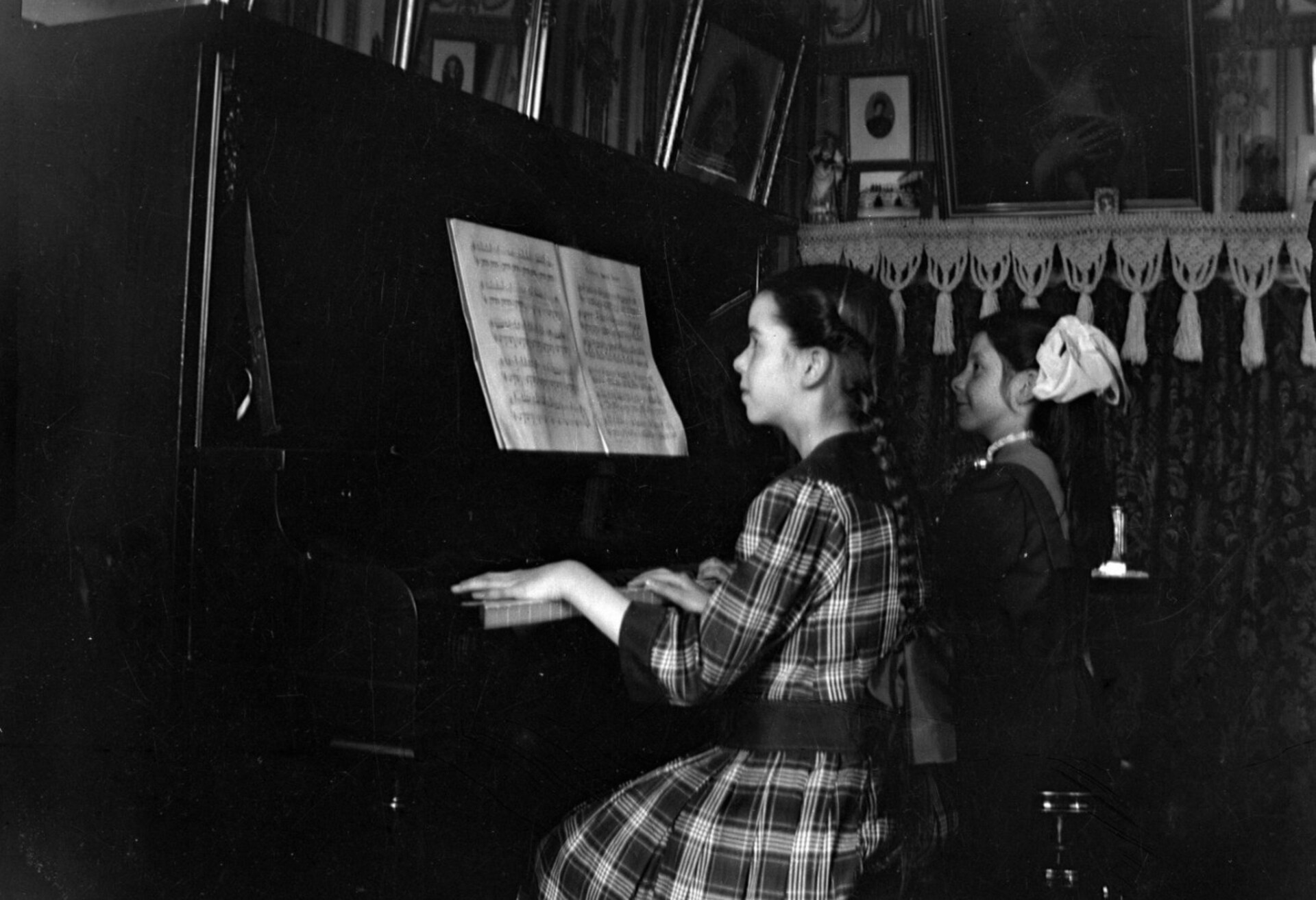 Deux jeunes filles sont assises au piano dans un chic salon de 1925. L'une d'elle déchiffre attentivement une partition placée devant elle, alors que l'autre jeune fille l'assiste en tournant les pages Deux jeunes filles sont assises au piano dans un chic salon de 1925. L'une d'elle déchiffre attentivement une partition placée devant elle, alors que l'autre jeune fille l'assiste en tournant les pages