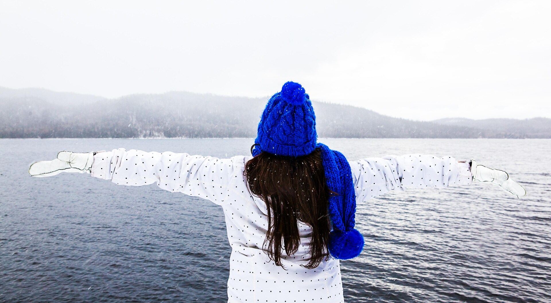 Valérie Jessica Laporte de dos bras écartés face à une étendue d'eau, vêtue d'un manteau et de mitaines blanches, avec une tuque et un foulard bleus.