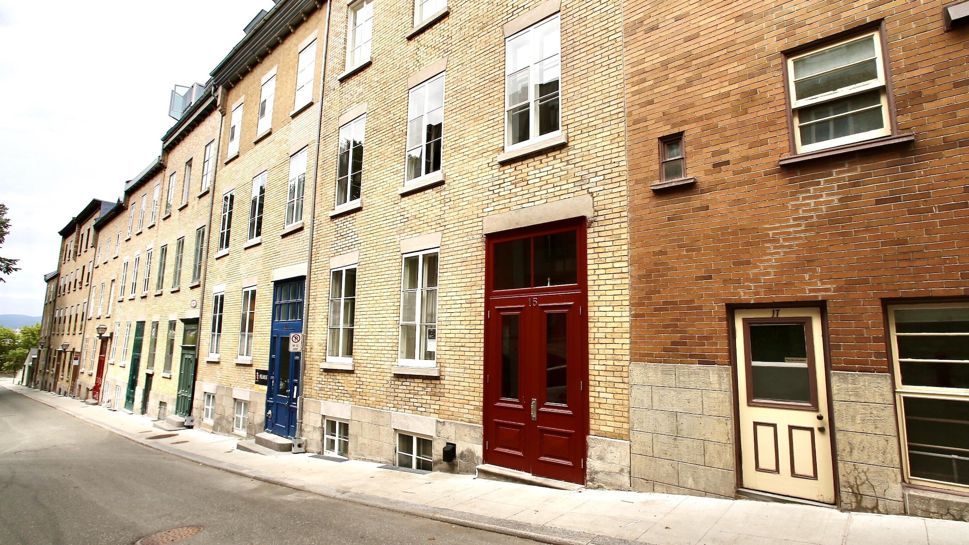 Une série de maisons mitoyennes descendent vers les arbres de la rue des Remparts, dans une enfilade de façades de briques jaunes et de portes aux couleurs vives. Une série de maisons mitoyennes descendent vers les arbres de la rue des Remparts, dans une enfilade de façades de briques jaunes et de portes aux couleurs vives.