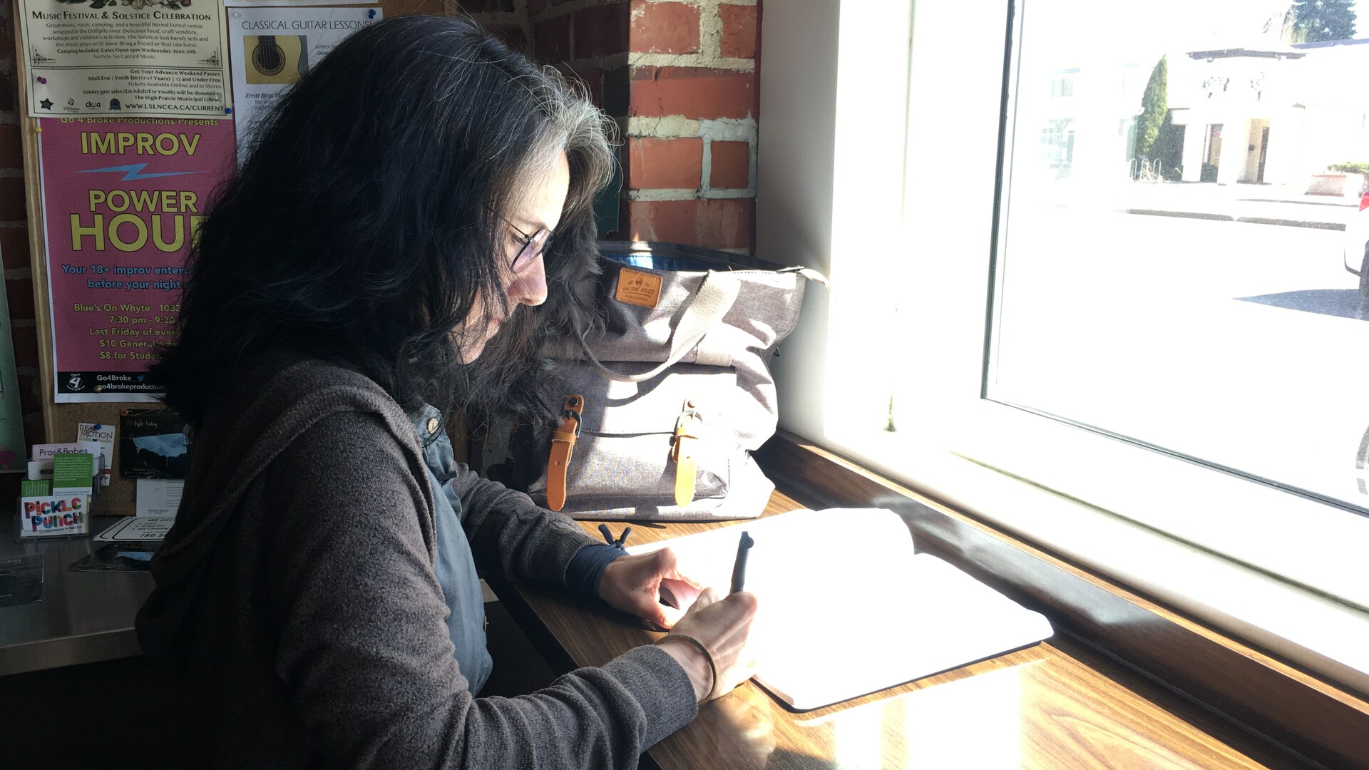 Portrait de profil d'une femme assise dans un café, devant une fenêtre, en train d'écrire èa la main dans un cahier.